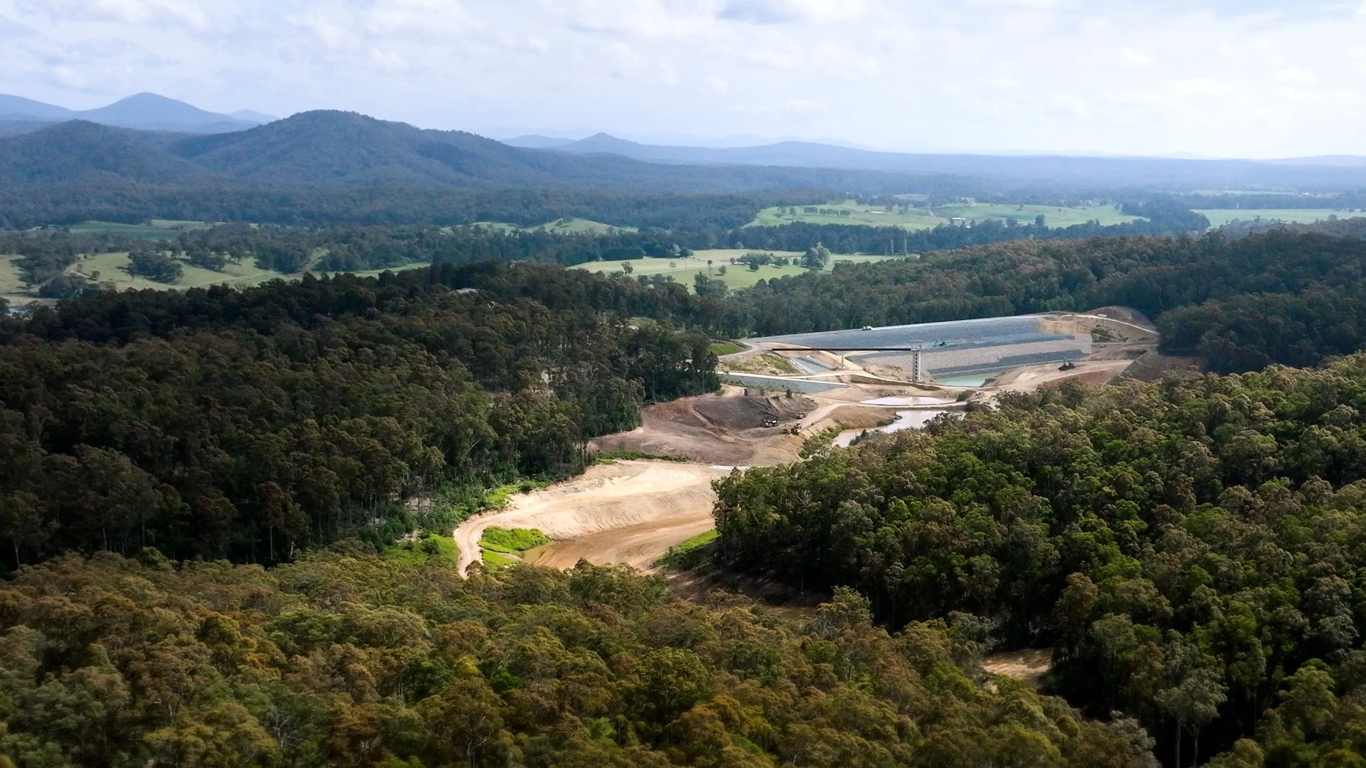 Aerial view over forested hills to the Eurobodalla Southern Storage Dam under construction, with farmland and ranges beyond